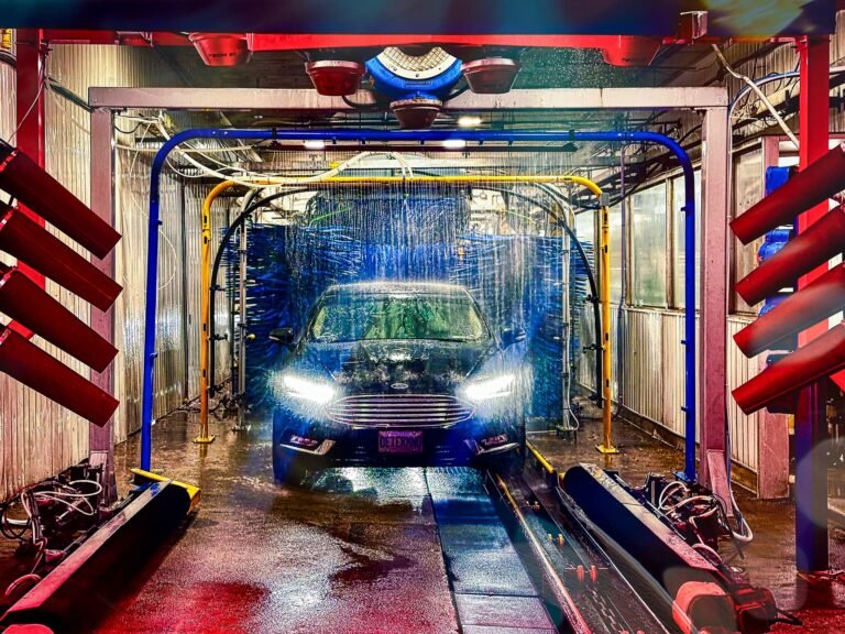 A sleek black car undergoing a fully automated car wash process, surrounded by vibrant red, yellow, and blue machinery, with water and soap jets actively cleaning the vehicle. The bright lighting highlights the advanced technology and spotless environment of the car wash facility.